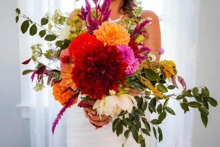 Braut hält einen Brautstrauß mit großer roter Tellerdahlie sowie orangefarbenen und rosa Balldahlien vor dem weißen Hochzeitskleid